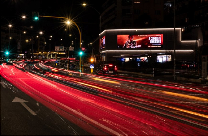 Schermo pubblicitario luminoso su edificio all’angolo di una strada notturna, con flusso continuo di auto e scie di luce.