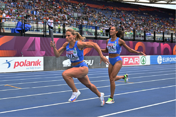 Atlete italiane in gara di staffetta su pista d’atletica, con pubblico sugli spalti e sponsor a bordo campo.