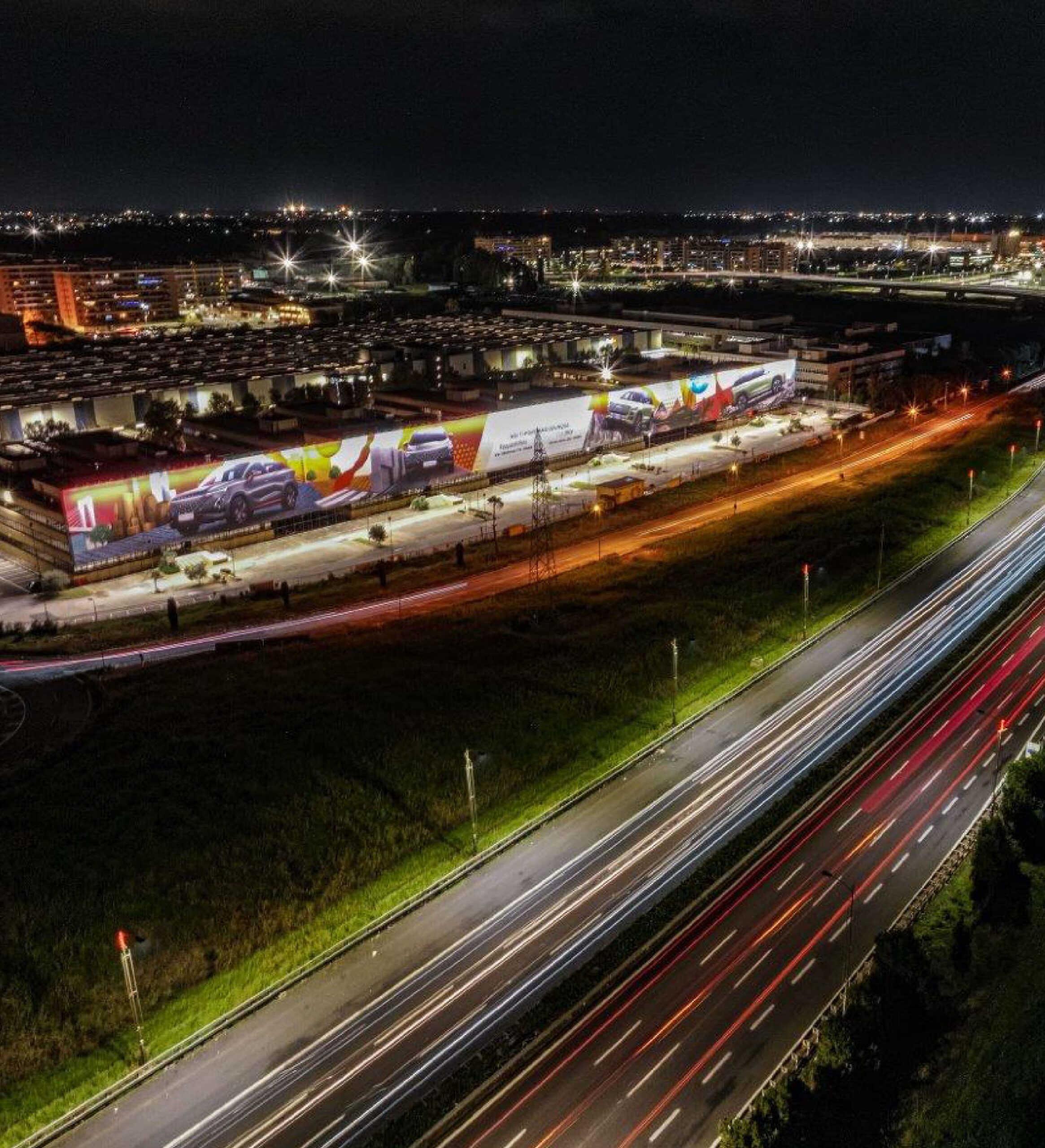 Vista notturna di una grande affissione pubblicitaria lungo un’area industriale, con autostrada illuminata e scie di traffico.