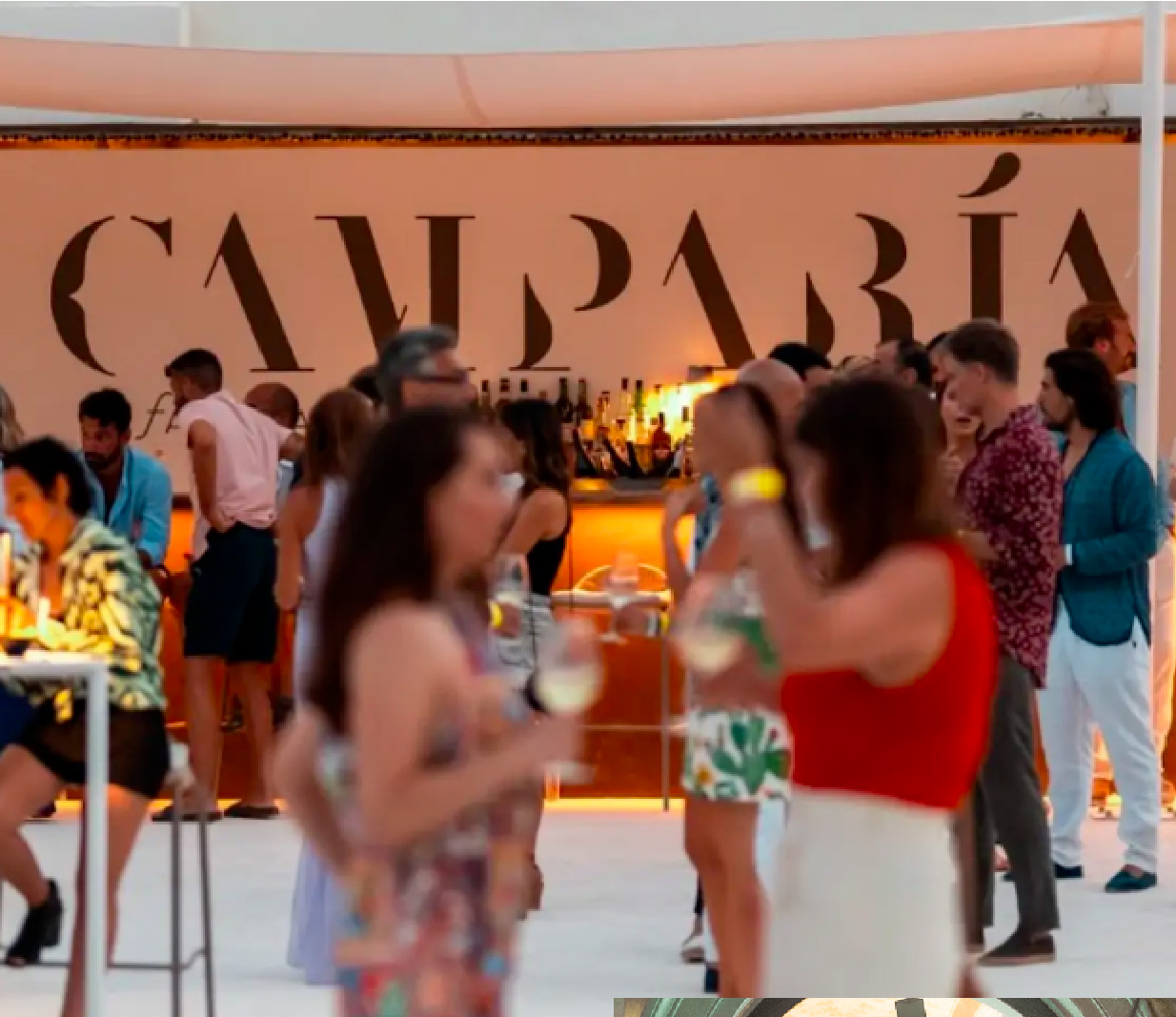 Persone partecipano a un evento serale davanti a un bancone bar illuminato con il logo Camparì sullo sfondo; gli ospiti, in abiti estivi, chiacchierano e sorseggiano drink in un’atmosfera vivace e conviviale