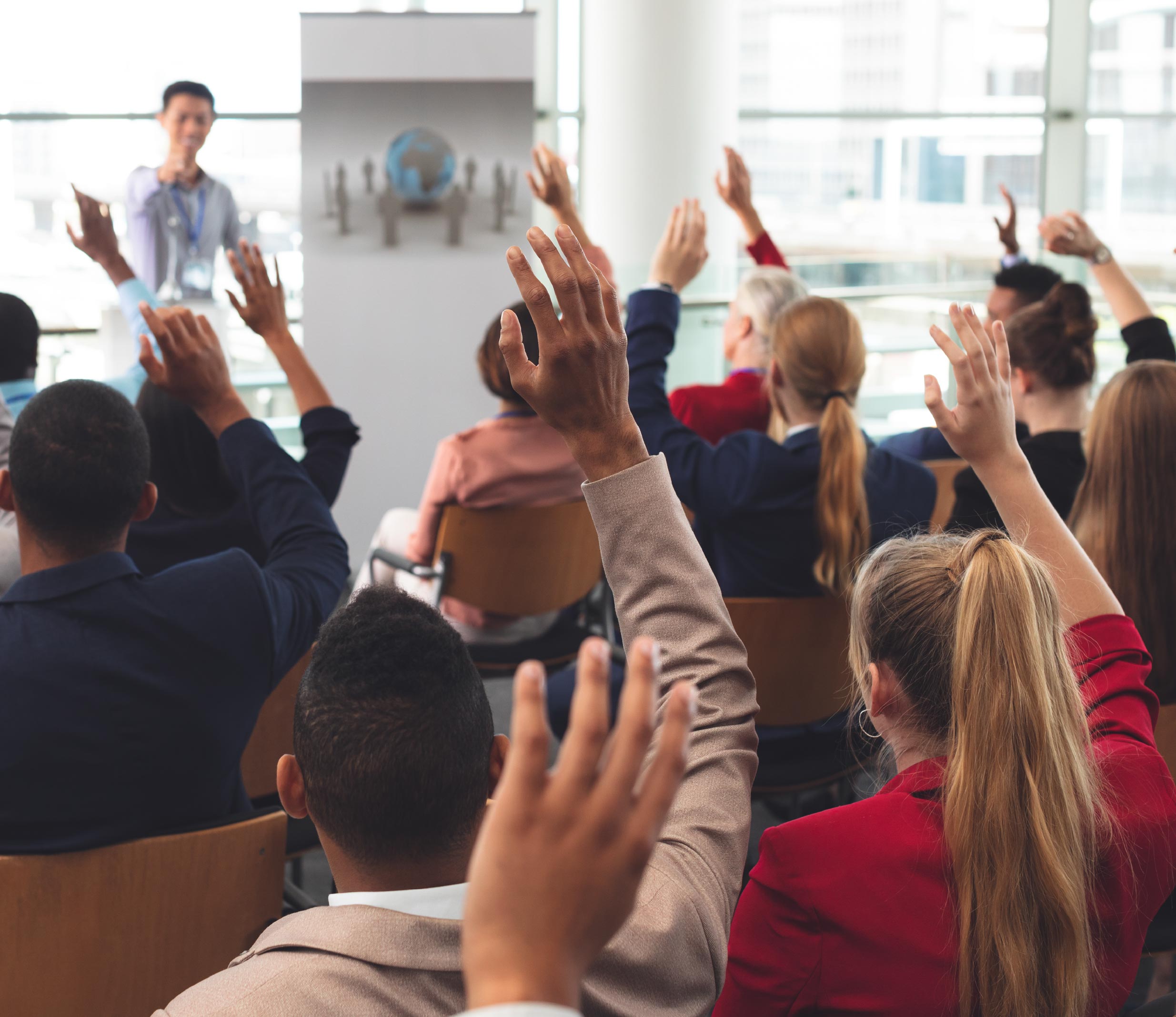 Partecipanti a una conferenza seduti in sala alzano la mano per intervenire, mentre un relatore parla davanti a un pubblico attento in un ambiente luminoso e professionale