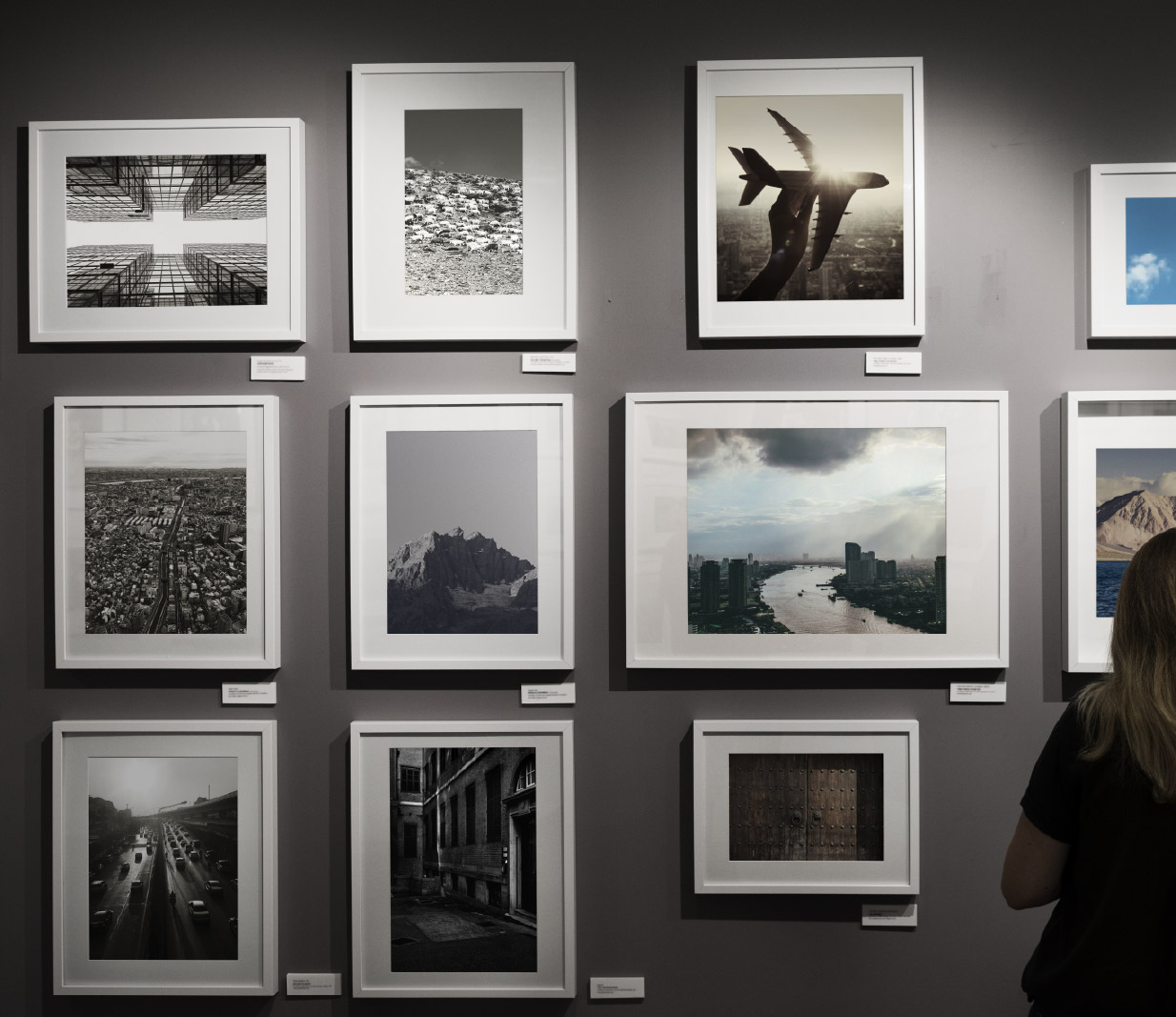 Parete di galleria con diverse fotografie incorniciate appese, che mostrano paesaggi urbani, natura e soggetti diversi, in un ambiente espositivo con illuminazione interna