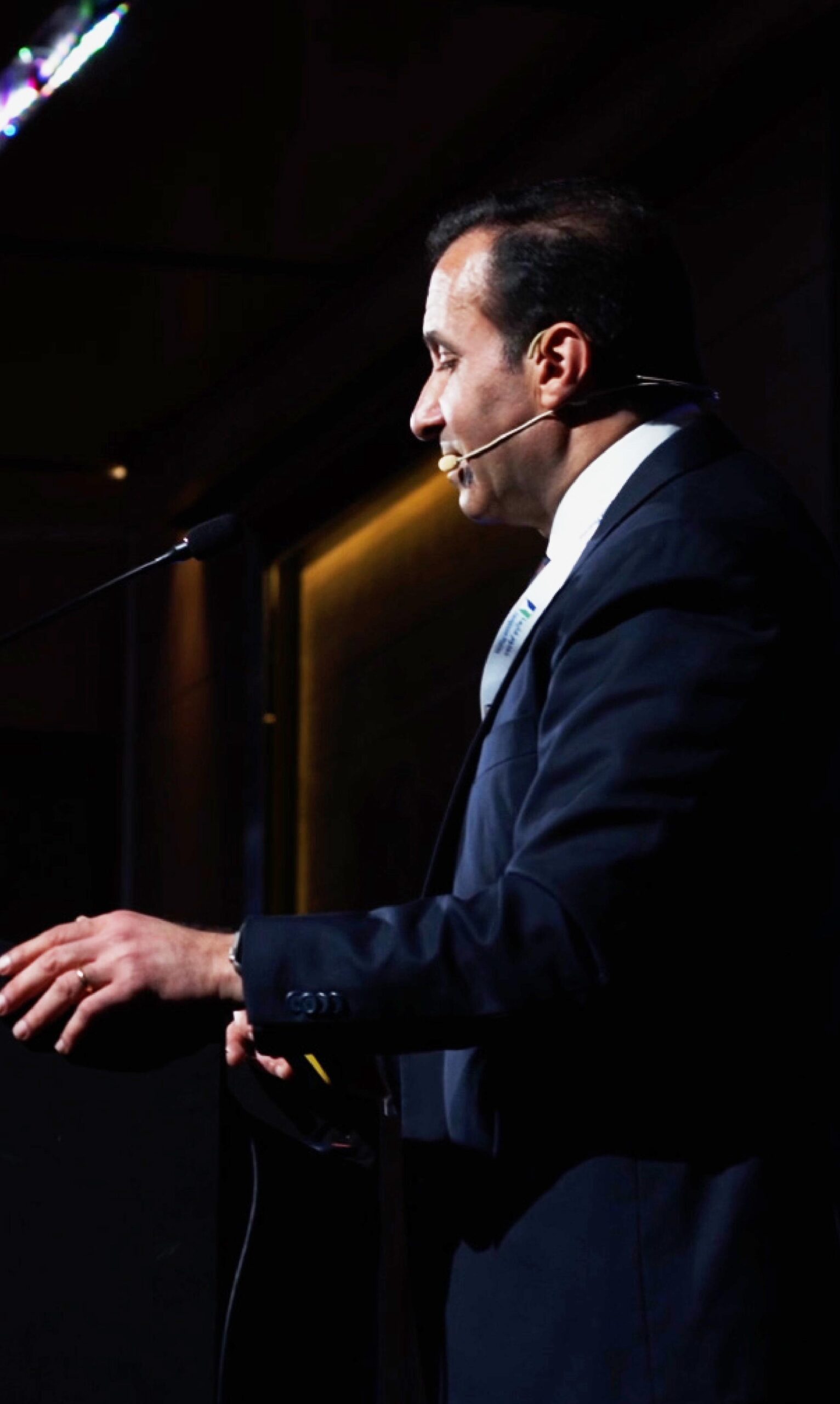 Relatore in abito formale parla a un pubblico durante un convegno, ripreso di profilo mentre utilizza un microfono headset e gesticola dal podio, illuminato da luci sceniche in un ambiente professionale