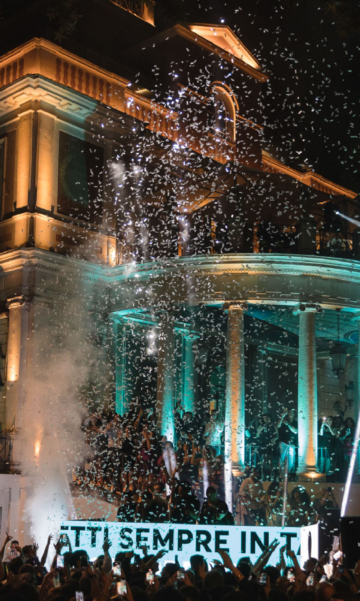 Evento pubblico serale davanti a un edificio storico illuminato con luci colorate, con una folla di persone in festa, coriandoli nell’aria e un’atmosfera di celebrazione
