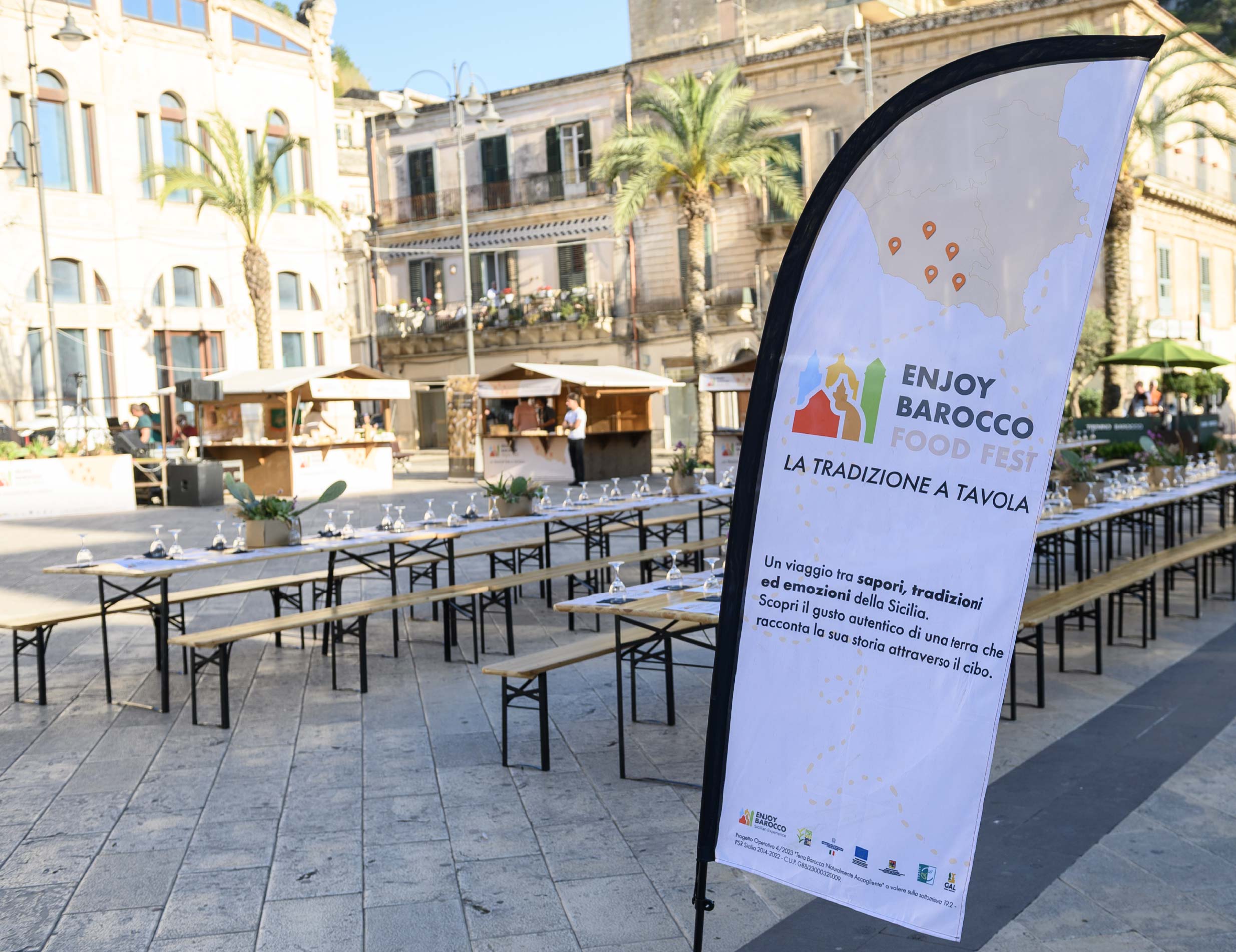 piazza di un centro città allestita da stand in legno, tavoli e panche apparecchiate e in primo piano lo striscione verticale di Enjoy Barocco