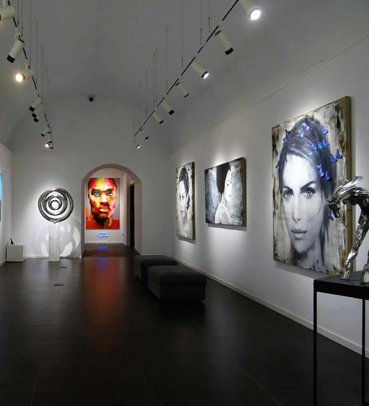 Galleria d’arte moderna con opere appese alle pareti, sculture e installazioni esposte sotto luci da soffitto in un ambiente bianco e luminoso, con quadri di volti stilizzati e un ritratto colorato sul fondo